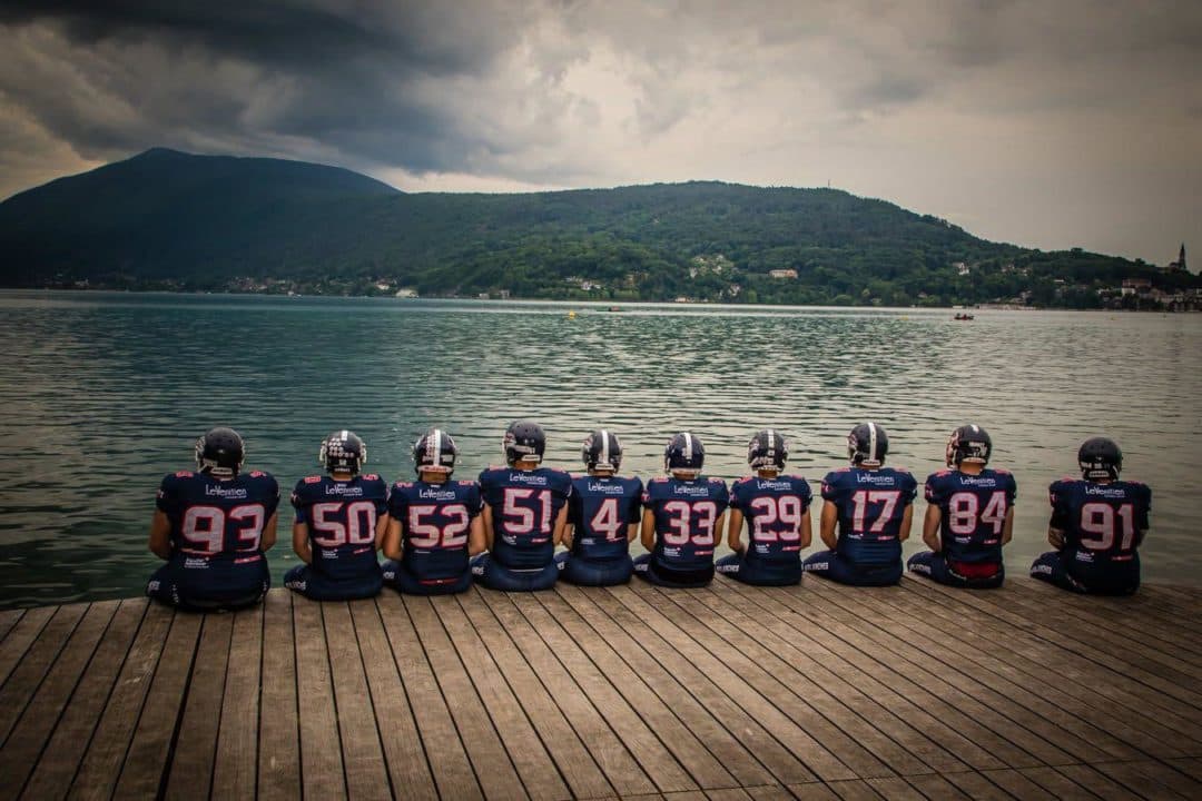 Dix joueurs des Avalanches assis de dos sur un ponton du lac d'Annecy, maillots numérotés face aux montagnes